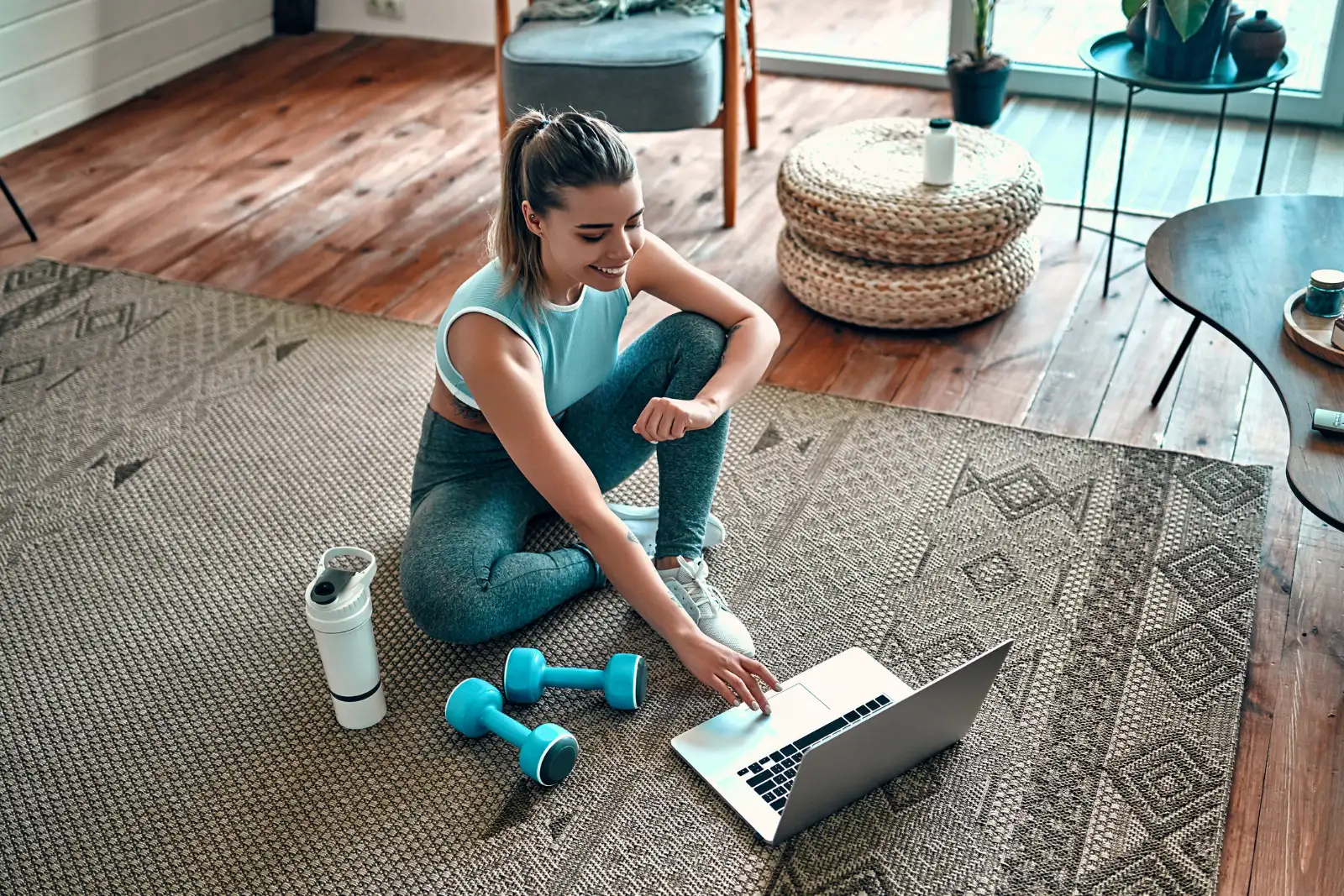 Online-Fitness: Flexibel trainieren von zu Hause aus Junge Frau im Sportoutfit sitzt mit Laptop, Hanteln und Trinkflasche im Wohnzimmer beim Online-Training.