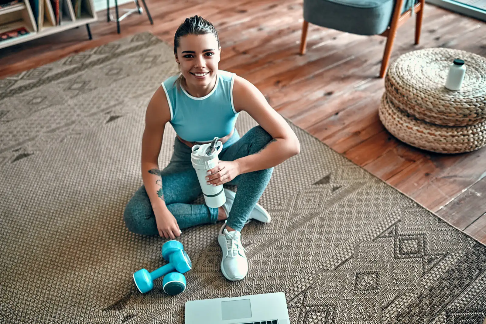 Zufrieden nach dem Training – Online-Fitness mit Erfolgserlebnis Sportlich gekleidete Frau sitzt nach dem Workout mit Trinkflasche und Hanteln auf dem Teppich, Laptop vor ihr.