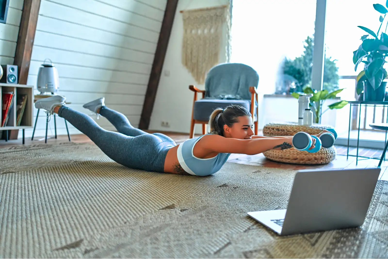 Starker Rücken – funktionelles Training mit Anleitung von zu Hause Frau in Sportkleidung führt Superman-Übung mit Kurzhanteln im Wohnzimmer vor dem Laptop aus.